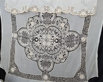 May include: A sheer white lace tablecloth with an intricate floral design. The tablecloth features a central medallion and a decorative border. The edges are scalloped and adorned with small floral accents. The background is black.