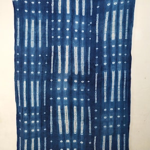 Puede incluir: Textil azul índigo con un patrón repetitivo de rayas verticales blancas, diamantes y puntos. La tela tiene una apariencia texturizada y un borde deshilachado. El diseño es simétrico y cubre toda la superficie del textil.