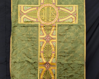 May include: Olive green liturgical vestment with a large embroidered cross design. The cross features gold, red, and yellow accents, with a central circular emblem. The vestment has a floral pattern and is trimmed with gold and pink.