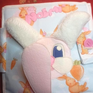 Può includere: Un libro di stoffa fatto a mano con un disegno di coniglietto. Il coniglietto ha caratteristiche rosa e bianche, con un occhio blu e un naso rosa. Il libro è azzurro e bianco, con illustrazioni di coniglietti e carote. Il nome "Noemi" è scritto in rosa.
