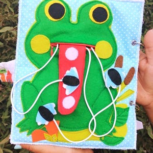 Puede incluir: Libro de actividades de fieltro hecho a mano con temática de rana. La portada presenta una rana verde con grandes ojos amarillos y una lengua roja. El libro tiene un fondo azul con lunares blancos y elementos interactivos.
