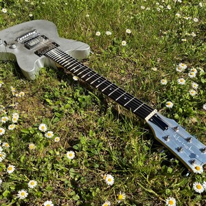 May include: A silver electric guitar with a black fretboard and strings rests in a field of green grass and white daisies. The guitar's headstock has the word "Hustler" printed on it.