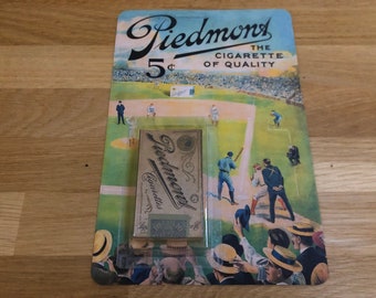May include: A vintage pack of Piedmont cigarettes, featuring a baseball scene with players and spectators. The pack is 5 cents and reads "The Cigarette of Quality".