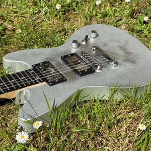 May include: A silver electric guitar with black pickups and a black fretboard rests in a bed of green grass and white daisies. The guitar has chrome hardware and is angled towards the viewer.