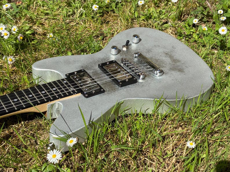 May include: A silver electric guitar with black pickups and a black fretboard rests in a bed of green grass and white daisies. The guitar has chrome hardware and is angled towards the viewer.