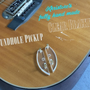 Puede incluir: Guitarra acústica con una pastilla de boca y soportes transparentes. La guitarra tiene un cuerpo de madera marrón claro con vetas visibles. El texto "Soundhole Pickup" y "Clear Brackets" son visibles en la guitarra. El texto "Kristore's fully hand made" también es visible.
