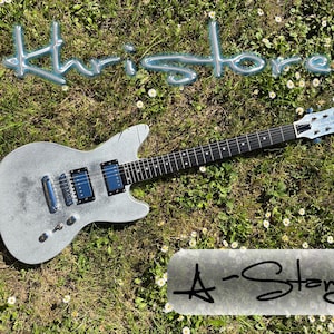 May include: A silver electric guitar with black and blue accents, resting on a bed of green grass and small white flowers. The word "Khristore" is written in a light blue, 3D font above the guitar. The text "A-Stong" is written below the guitar.