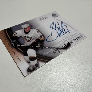 May include: A hockey trading card featuring Sidney Crosby in a Pittsburgh Penguins uniform. The card has an autograph and the text "sign of the times". The card is on a white surface.