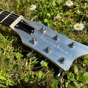 May include: Close-up of a silver guitar headstock with the name "Khristore" in script. The guitar strings are visible, along with tuning pegs. The headstock is set against a backdrop of green grass and small white and yellow flowers.