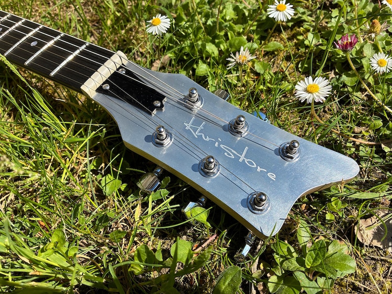 May include: Close-up of a silver guitar headstock with the name "Khristore" in script. The guitar strings are visible, along with tuning pegs. The headstock is set against a backdrop of green grass and small white and yellow flowers.