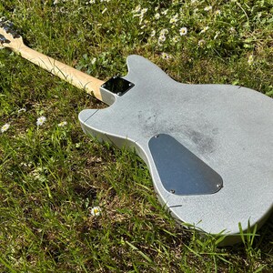 May include: A silver electric guitar with a light wood neck and headstock lies in green grass with small white flowers. The guitar's body has a metallic finish, and a black plate covers the back.
