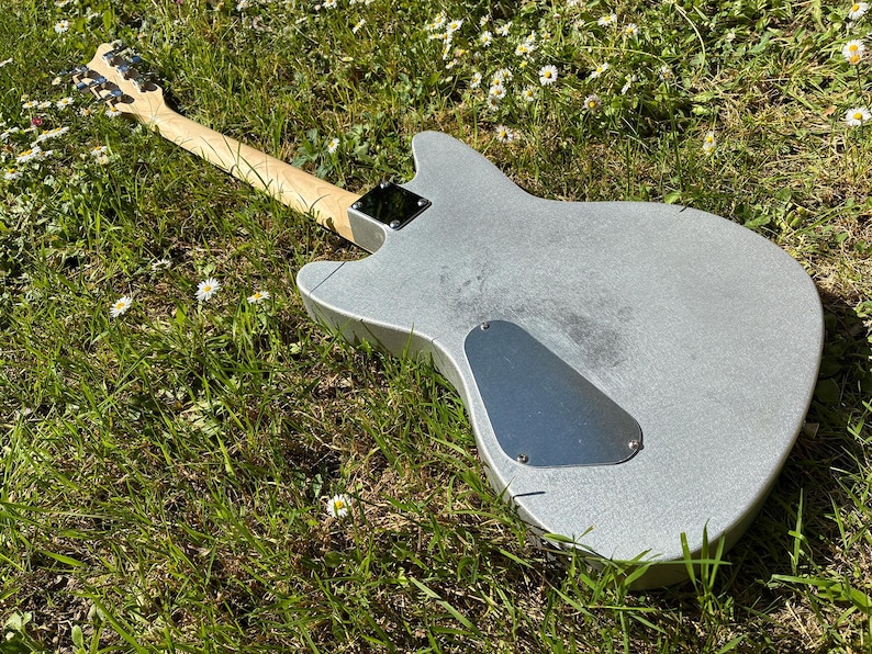 May include: A silver electric guitar with a light wood neck and headstock lies in green grass with small white flowers. The guitar's body has a metallic finish, and a black plate covers the back.