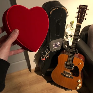 Puede incluir: Una caja de terciopelo rojo en forma de corazón junto a una guitarra acústica marrón en una funda negra. La guitarra tiene una golpeador blanco y una sola perilla en la parte inferior.