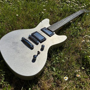 May include: A silver electric guitar with black pickups and a black fretboard rests on a bed of green grass and white flowers. The guitar has a metallic finish and chrome hardware.