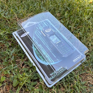 May include: A Samsung Matrix Reloaded edition mobile phone in its original clear plastic blister packaging. The black phone features a numeric keypad and screen, with the Matrix Reloaded logo and digital rain graphics printed on the backing card.