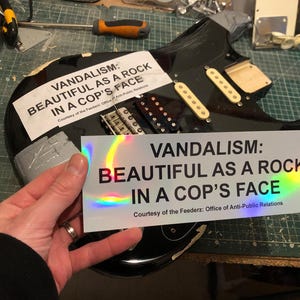 Op de afbeelding: Een zwarte elektrische gitaar met een sticker waarop staat "Vandalism: Beautiful as a rock in a cop's face. Courtesy of the Feederz: Office of Anti-Public Relations."