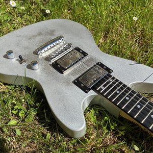 May include: A silver electric guitar with a black fretboard and chrome hardware rests on a bed of green grass. The guitar features two pickups, control knobs, and a bridge. The instrument is designed for playing music.