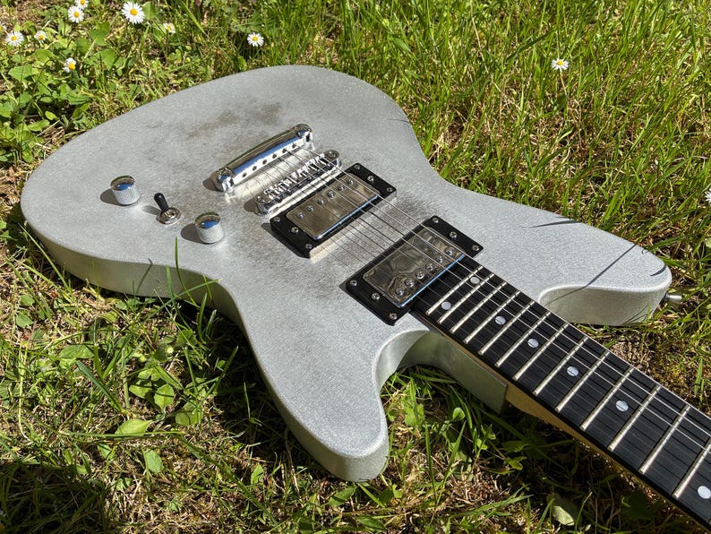 May include: A silver electric guitar with a black fretboard and chrome hardware rests on a bed of green grass. The guitar features two pickups, control knobs, and a bridge. The instrument is designed for playing music.