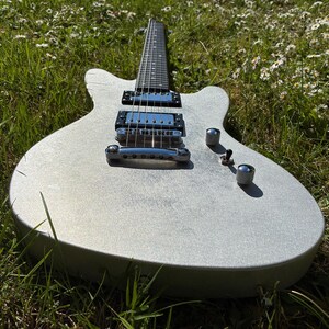 May include: A silver electric guitar with a double-cutaway body rests in a field of green grass and white flowers. The guitar has chrome hardware, including pickups, knobs, and a bridge. The fretboard is black, and the body has a textured finish.