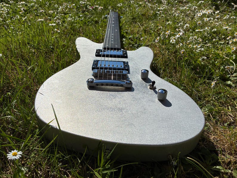 May include: A silver electric guitar with a double-cutaway body rests in a field of green grass and white flowers. The guitar has chrome hardware, including pickups, knobs, and a bridge. The fretboard is black, and the body has a textured finish.