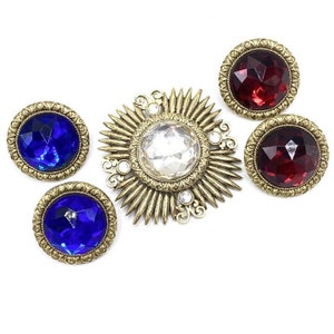 Może przedstawiać: Trzy złote broszki z fasetowanymi szklanymi kamieniami. Jedna broszka ma przezroczysty kamień w kształcie słońca. Dwie pozostałe broszki mają czerwone i niebieskie kamienie.