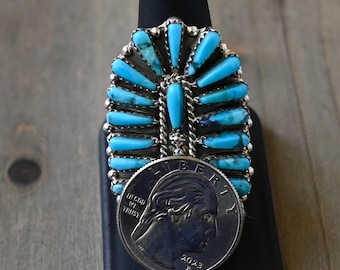 May include: A silver ring with a turquoise stone design. The ring features multiple turquoise stones arranged in a fan shape. A US quarter is placed next to the ring for size comparison.