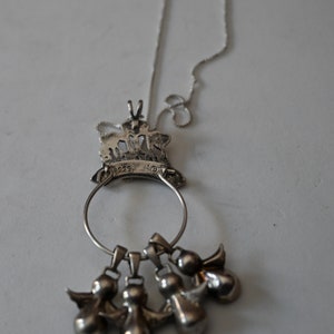 May include: A silver necklace with a pendant featuring a crown with angels. The pendant is attached to a silver chain and has five small angel charms hanging from it.