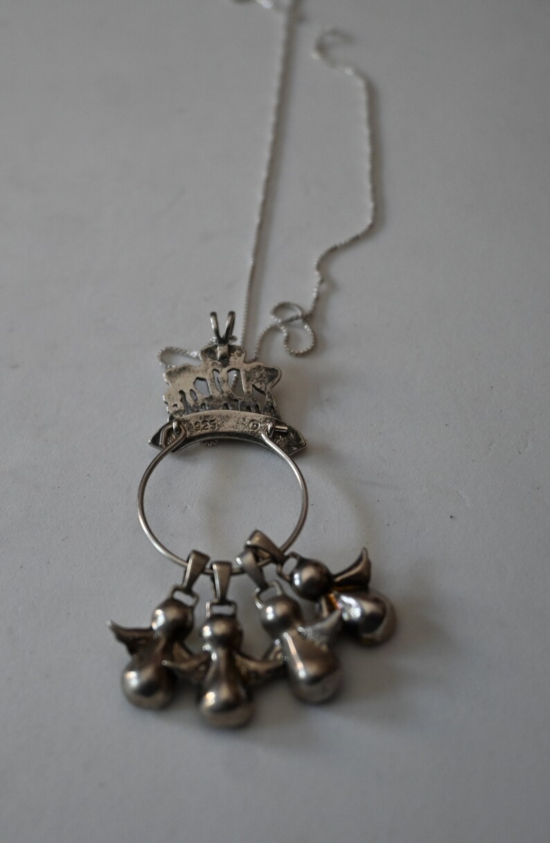 May include: A silver necklace with a pendant featuring a crown with angels. The pendant is attached to a silver chain and has five small angel charms hanging from it.