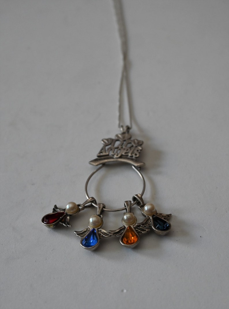 May include: A silver necklace with a charm that says "Our Angels" and four dangling angel charms with red, blue, orange, and white gemstones.
