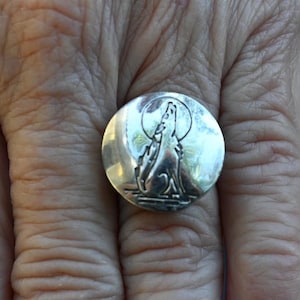 May include: A silver-toned round ring with a detailed relief of a howling wolf under a full moon. The ring is set against a background of skin, highlighting the intricate design and metallic sheen. The wolf is depicted in a stylized, artistic manner.