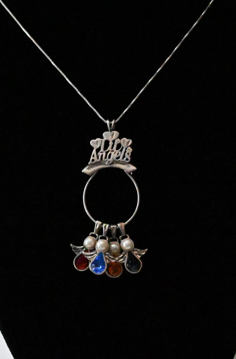 May include: Silver necklace with a charm that says "Lil Angels" and has a circle with four angel charms hanging from it. Each angel charm has a different coloured gemstone.