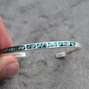 Op de afbeelding: Een zilveren manchetarmband met turkoois inlegwerk. De open armband heeft rechthoekige turkooizen stenen gezet in een zilveren band. Een sieraad.