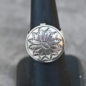 May include: A silver ring with a large, circular face featuring a detailed, symmetrical design. The design resembles a stylized flower or sunburst. The ring is displayed on a black stand against a neutral background.