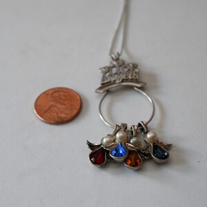 May include: Silver necklace with a charm that says "Los Angeles" and four dangling angel charms with red, blue, orange, and blue gemstones.