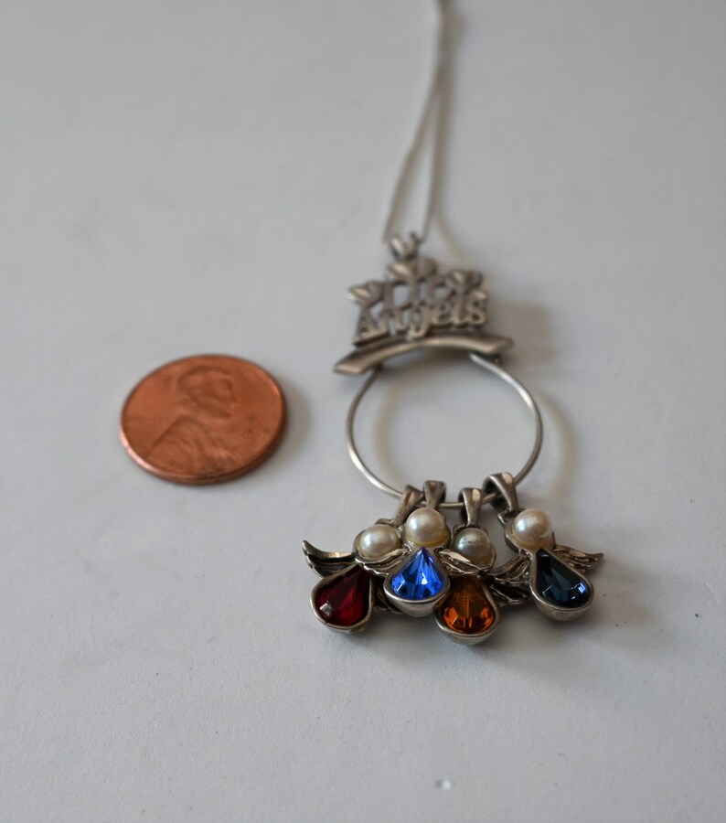 May include: Silver necklace with a charm that says "Los Angeles" and four dangling angel charms with red, blue, orange, and blue gemstones.