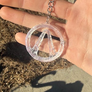 May include: Clear, circular keychain featuring a stylized "A" within a circle. The keychain is attached to a silver metal ring and chain. The design is transparent, allowing light to pass through. The keychain is held in a hand.