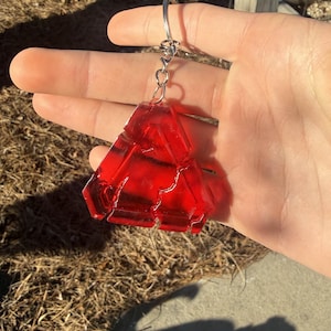 May include: A red, geometric-shaped keychain with a silver keyring. The keychain is made of a translucent material, allowing light to pass through. The design features a series of connected, angular shapes.