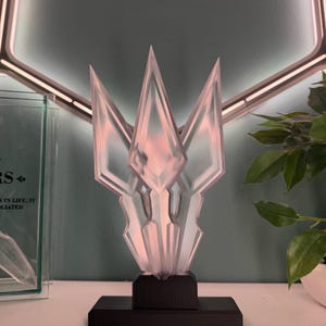 May include: A clear glass sculpture with three points, resembling a crystal or a crown, on a black base. The sculpture is illuminated from within, creating a soft, pink glow.