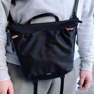 May include: Black crossbody bag with a textured, perforated design. The bag features a top handle, adjustable shoulder strap, and orange zipper pulls. The bag is worn across the body.