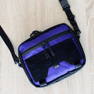 May include: A black and purple crossbody bag with a black adjustable strap and buckle. The bag features a black velcro patch and a zippered pocket. The text "11.58" is visible on the bag.