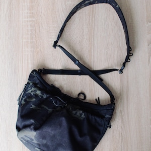 May include: Black camouflage crossbody bag with an adjustable strap. The bag features a drawstring closure and a small zipper pocket. The strap and bag are made of a durable, water-resistant material. The bag is designed for everyday use.