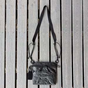 May include: A gray crossbody bag with a black adjustable strap and a small black tag. The bag has a zippered main compartment and a front pocket. The bag is hanging against a gray wooden background.