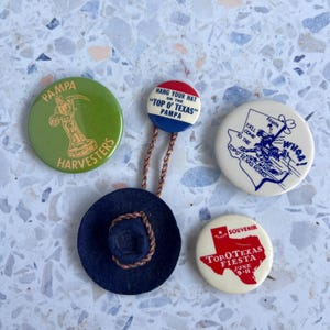 May include: A collection of vintage souvenir pins and a small hat. The pins feature text and graphics related to Pampa, Texas, including a rodeo, a fiesta, and the phrase "Top O' Texas." The hat is dark blue with a brown rope trim.