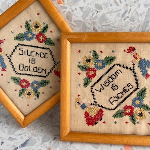 May include: Two framed cross-stitch samplers. One sampler reads "Silence is Golden" and the other reads "Wisdom is Riches". Both samplers feature floral and figure designs in red, blue, and green, set against a beige background, within light brown wooden frames.