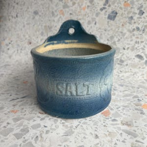 May include: A blue and white ceramic salt crock with a hanging loop. The crock has a faded blue glaze and the word "SALT" is embossed on the front.