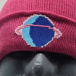 May include: A pink knit beanie with a blue and white pixelated design of a planet with a ring around it.