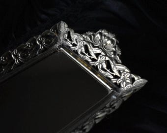 Puede incluir: Una bandeja de plata con un borde adornado y decorativo. La bandeja es rectangular y tiene una superficie reflectante.