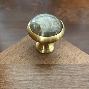 May include: A brass cabinet knob with a round, light gray stone top. The stone has a swirling pattern.