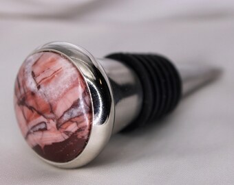 May include: A decorative wine bottle stopper with a polished, oval-shaped stone top. The stone features a mix of red, brown, and white veining, set in a silver-colored metal casing. The stopper has a black rubber seal and a tapered metal base.
