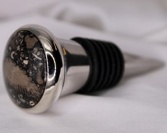 May include: A decorative wine bottle stopper with a stainless steel base and a black rubber seal. The stopper's top features a polished, round stone with a mix of black, white, and tan patterns. The design is elegant and suitable for wine enthusiasts.
